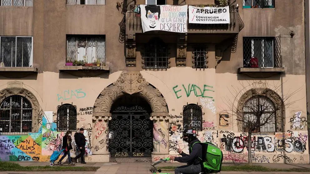 Un repartidor de una aplicación móvil y una pareja pasan frente a un edificio lleno de pintadas en referencias a las protestas, en Santiago de Chile.     FUENTE- EFE